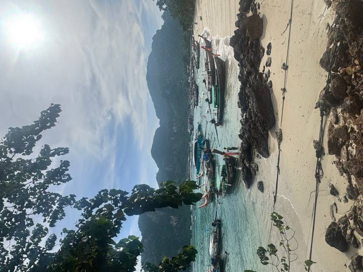 Long-tail boats anchored in turquoise water off a small sandy beach with jungle-covered mountains in the background.