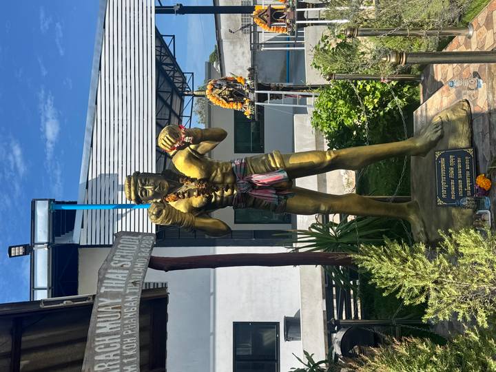 A golden Muay Thai boxer statue adorned with flower garlands stands outside a training school.