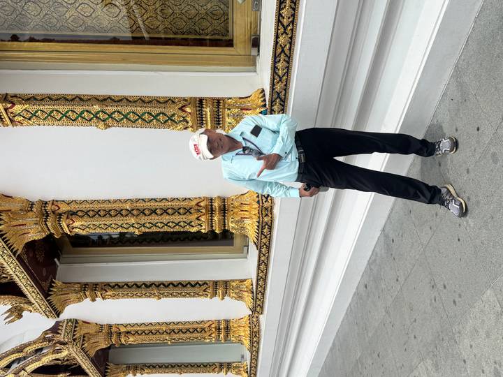 A smiling tourist poses in front of an intricately decorated Thai temple facade featuring gold and green motifs.