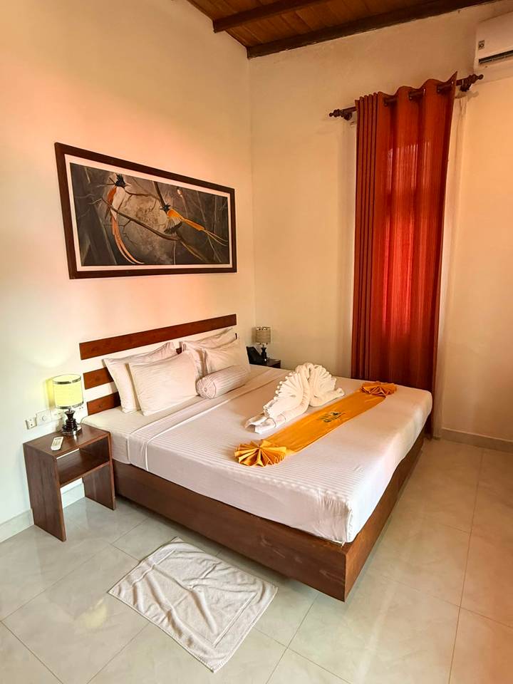 A neat hotel room with a large bed, crisp white linens and a decorative towel swan, accented by warm lighting and a framed bird artwork above the headboard.