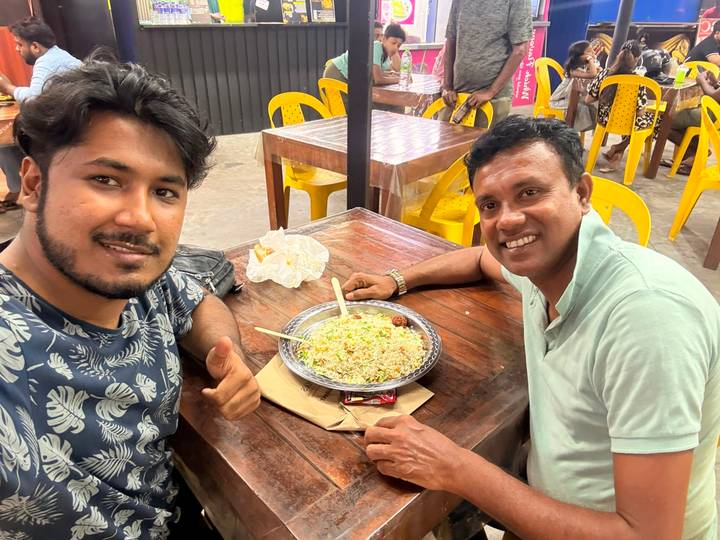 Two smiling men seated at a wooden table giving a thumbs-up while enjoying a plate of fried rice in a casual eatery.