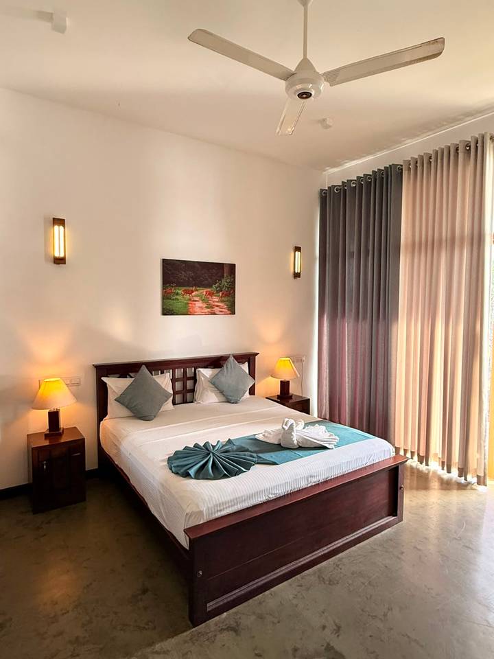 Bright, tidy hotel room featuring a double bed with teal accents, bedside lamps and tall sheer curtains letting in daylight.