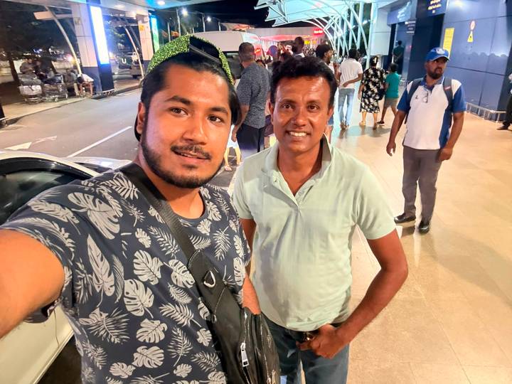 A selfie of two men on a lively city street at night, with pedestrians and lights in the background.