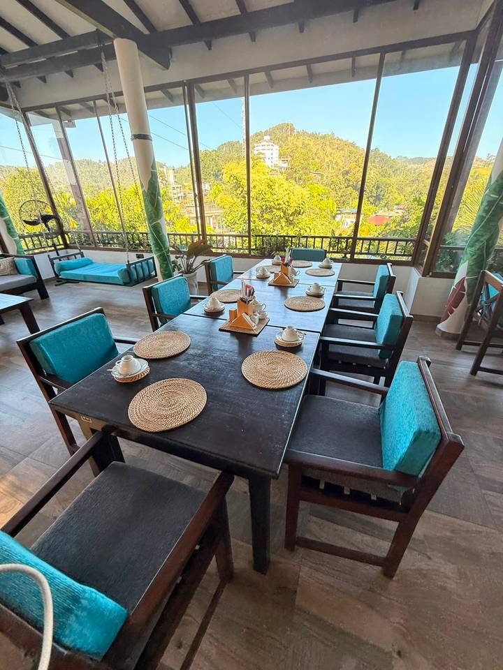 Open-air dining area with a long dark wooden table, turquoise cushions and views of greenery outside.