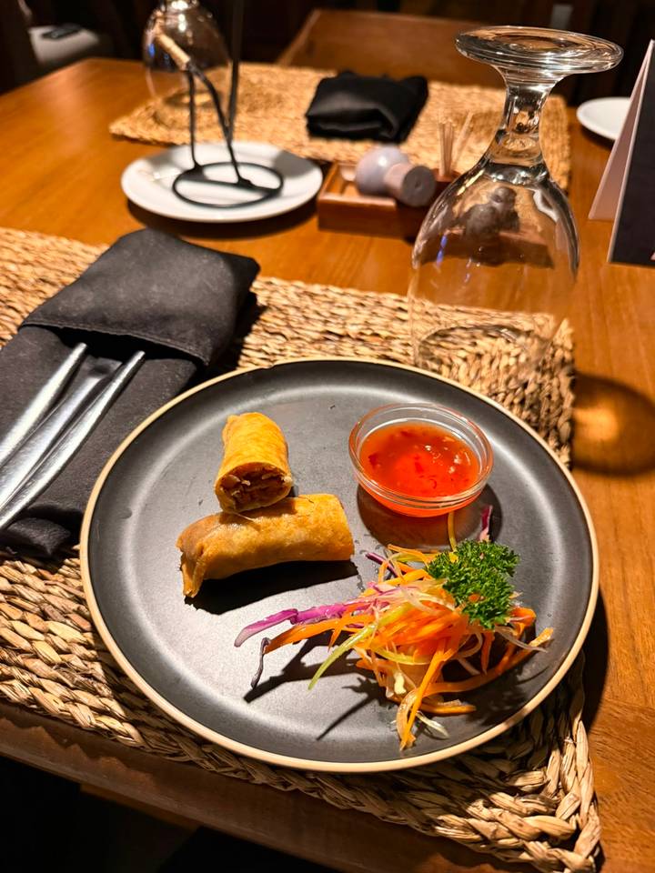 A black plate with two crispy spring rolls, sweet chili dipping sauce and vegetable garnish on a wooden table.