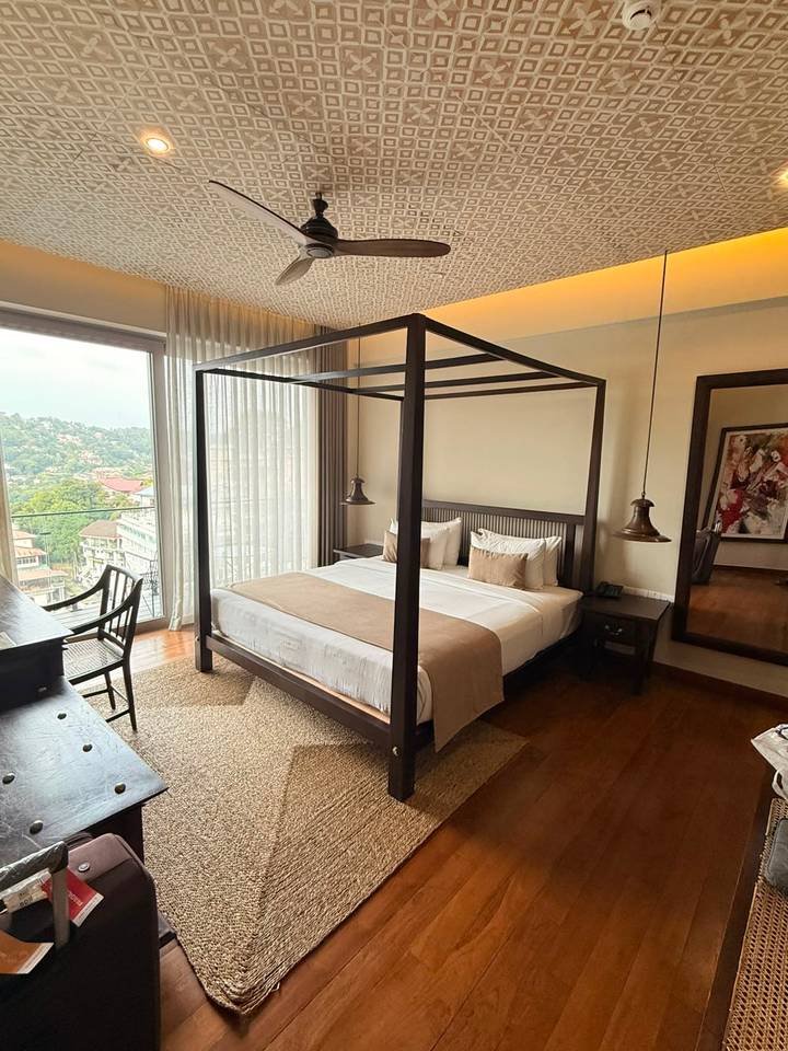 Elegant four-poster bed in a modern room with floor-to-ceiling windows overlooking a green hillside cityscape.