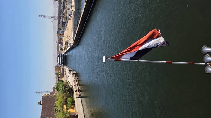 Egyptian flag atop a boat sails toward a narrow lock on a broad river under a clear sky.