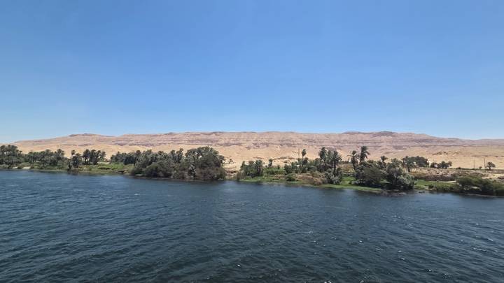 Blue waters of the Nile contrast with palm-lined banks and arid desert hills beneath a cloudless sky.