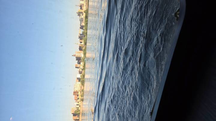 View across a wide river toward a modern city skyline under a clear blue sky, seen from a low vantage near the water.