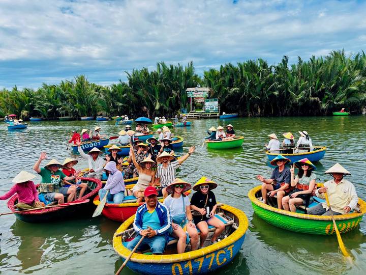 Dozens of travelers wearing conical hats sit in round basket boats on a calm tropical river lined with dense palms.