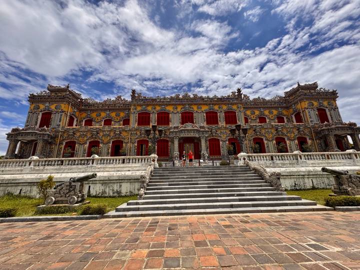 An ornate imperial-era building with bright red shutters and intricate mosaic details stands under a vivid blue sky.