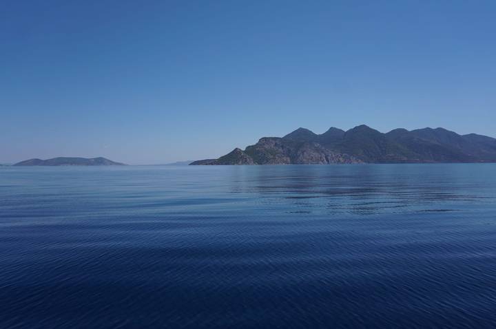 Glass-calm blue sea reflecting distant rugged islands under a clear midday sky.