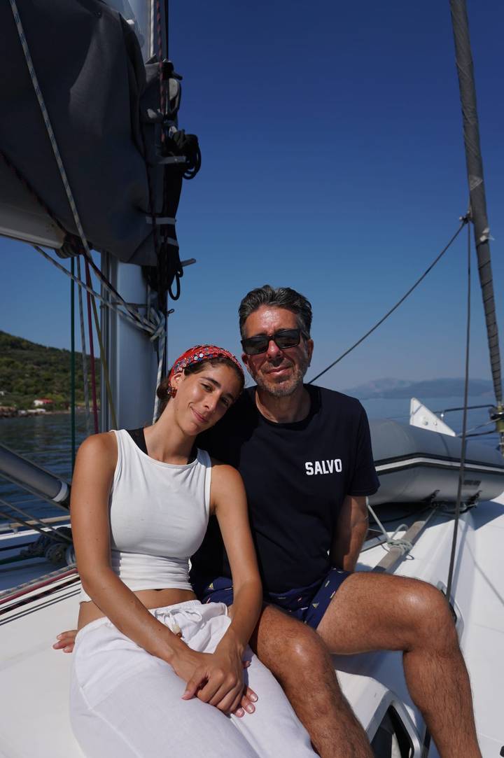 Close-up of two smiling people seated on a sailboat with calm sea and inflatable dinghy behind.