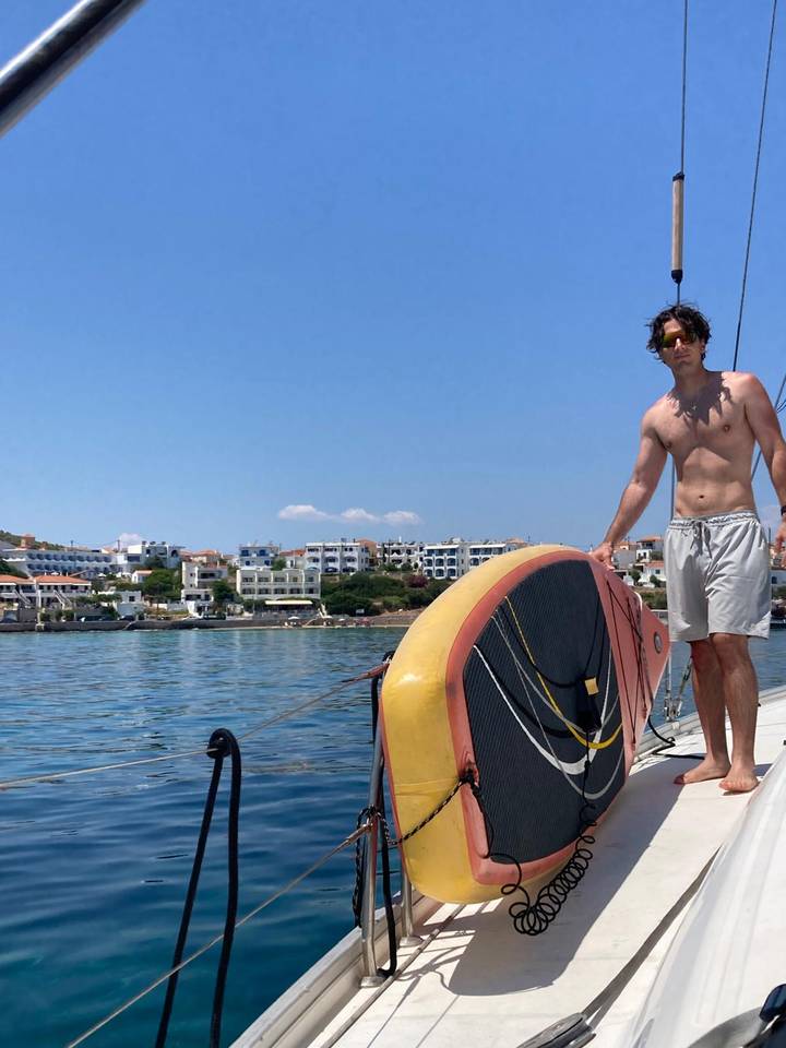 Young man on deck of sailboat holding a paddleboard with white seaside town in background.