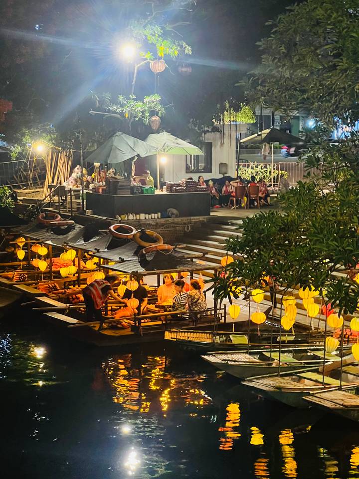 Night view of lantern-lit boats and riverside dining decks along a canal with people enjoying the evening.