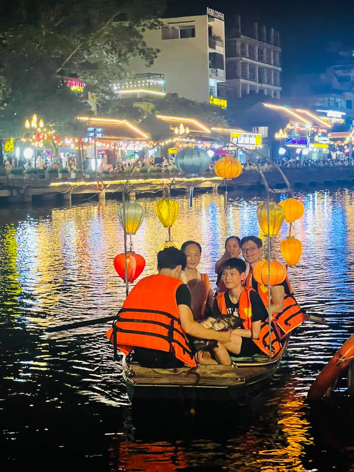 Tourists wearing life vests sit in a lantern-decorated boat on a shimmering river at night, city lights behind.
