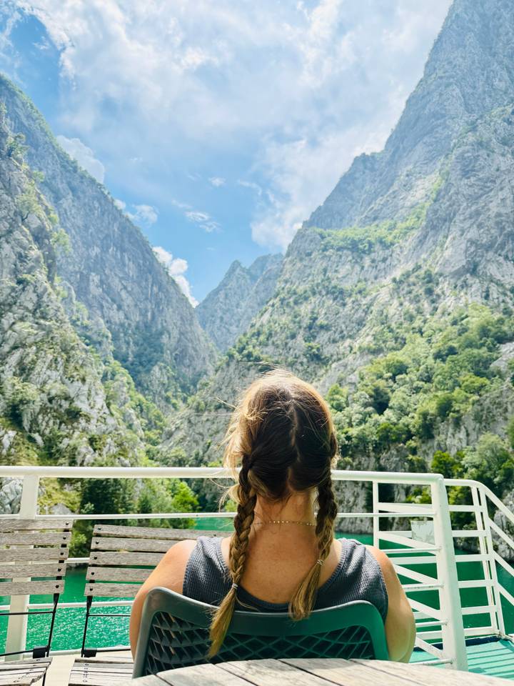 From behind, a traveler looks up at towering canyon walls thick with greenery while cruising on a boat.