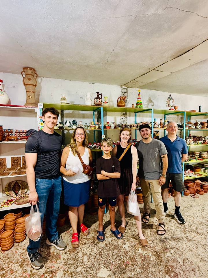 A small group of travelers poses inside a colorful pottery and souvenir shop filled with shelves of handmade items.