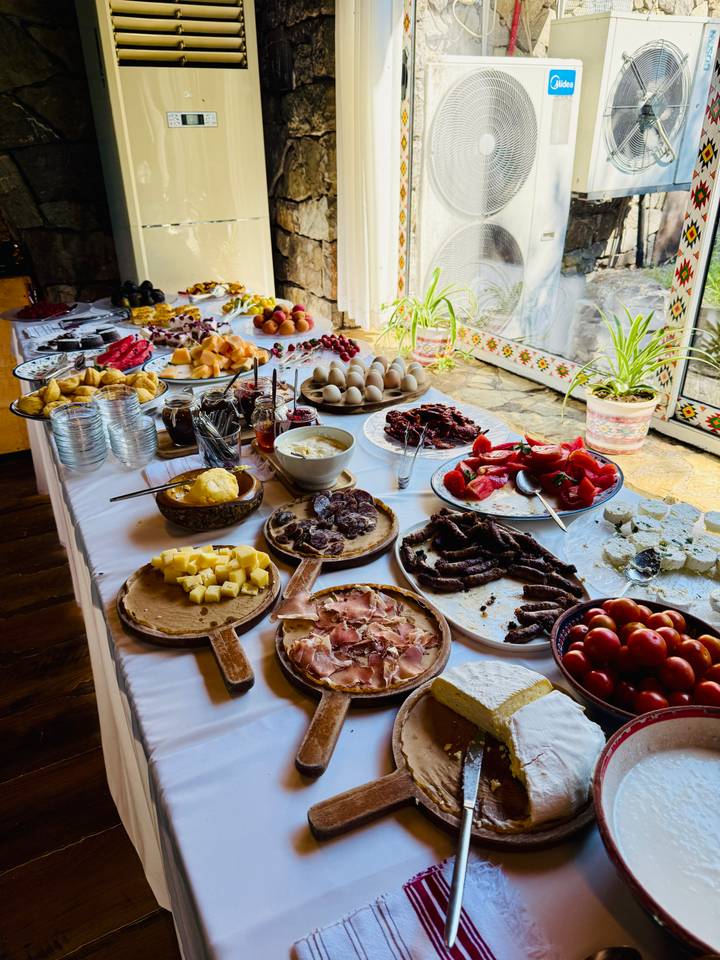 A long buffet table is covered with a rustic spread of local cheeses, meats, fruits and breads, lit by daylight from a window.