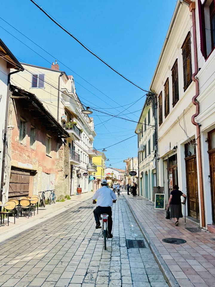 A narrow pedestrian street lined with pastel buildings and overhead cables shows locals strolling on a bright sunny day.
