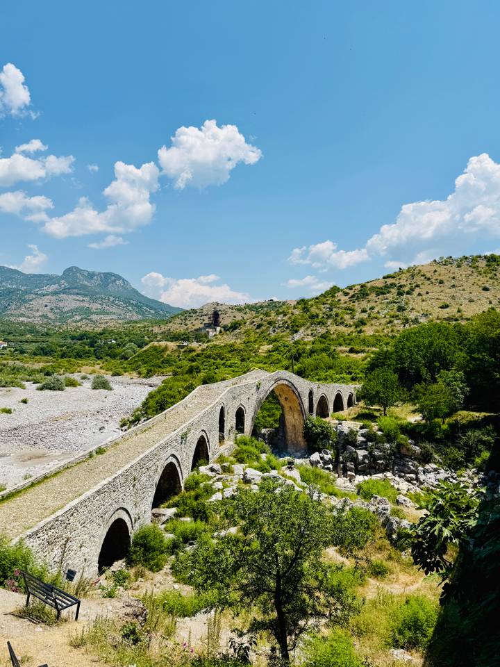 A historic multi-arched stone bridge spans a dry riverbed amid green hills and distant mountains under a bright sky.