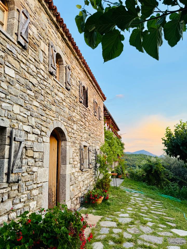 A rustic stone building with wooden shutters is bathed in the warm colors of sunset, with tree-covered hills beyond.