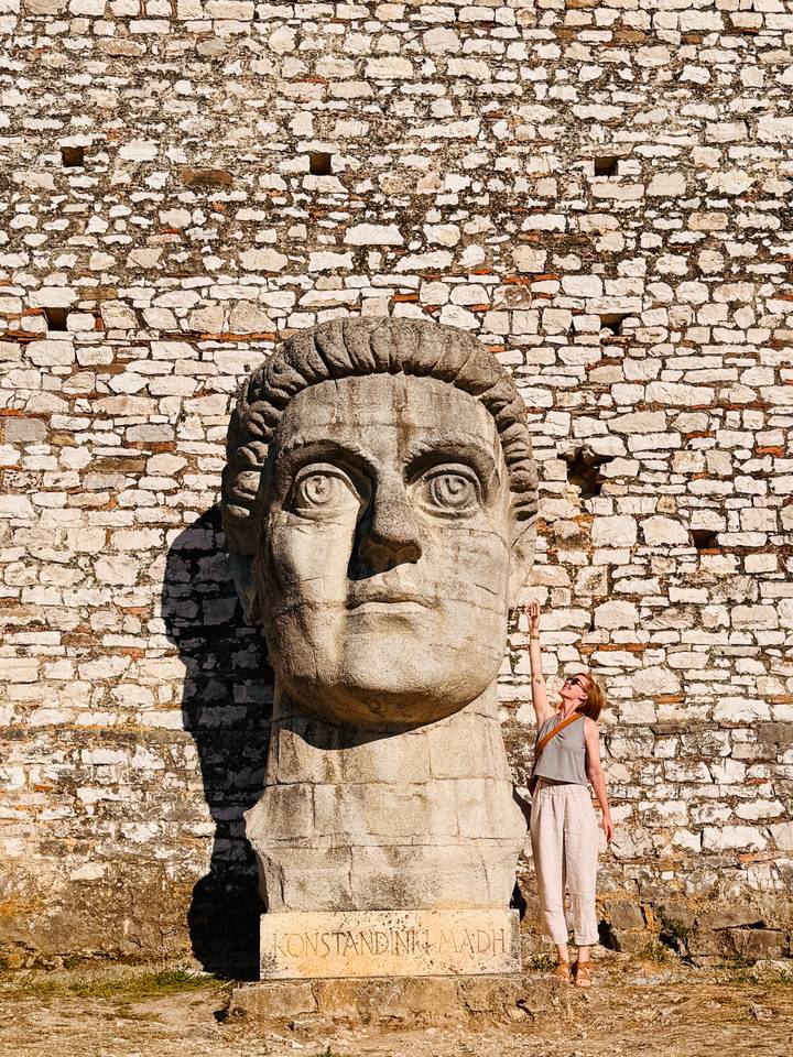 A woman playfully reaches up toward a gigantic stone head sculpture set against a historic brick wall.