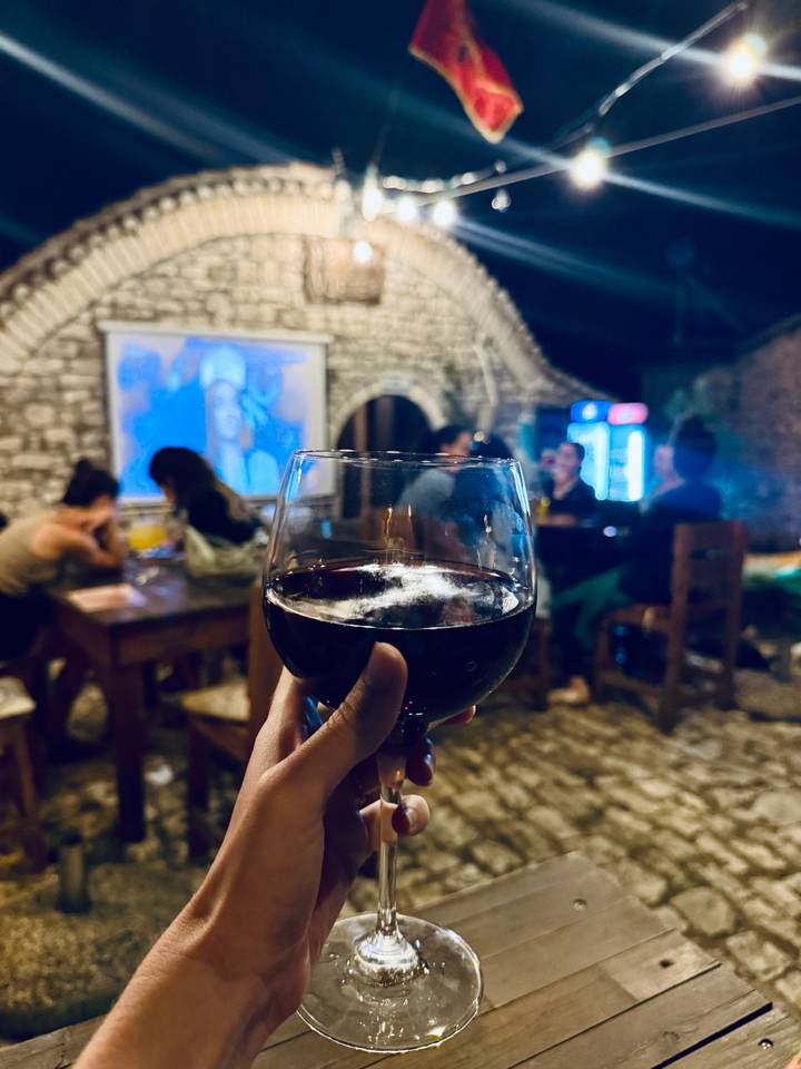 Close-up of a hand holding a glass of red wine inside a cozy stone-walled tavern with friends chatting in the background