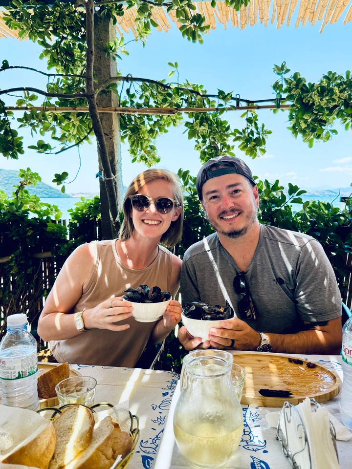 Smiling couple in sunglasses holding bowls of fresh mussels at a shaded waterfront restaurant