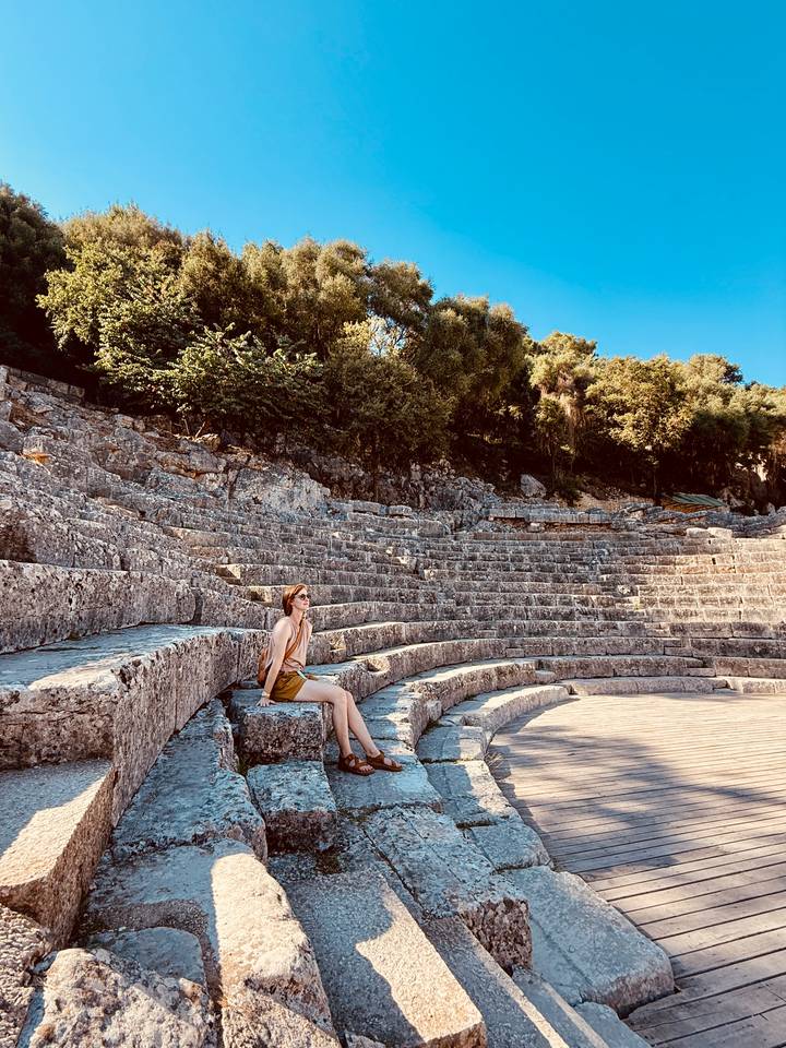 Woman resting on ancient stone tiers of a sun-drenched amphitheater surrounded by trees