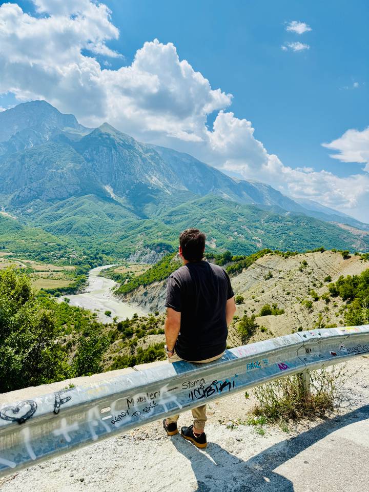 Man gazing over a winding river valley toward towering green mountains under scattered clouds