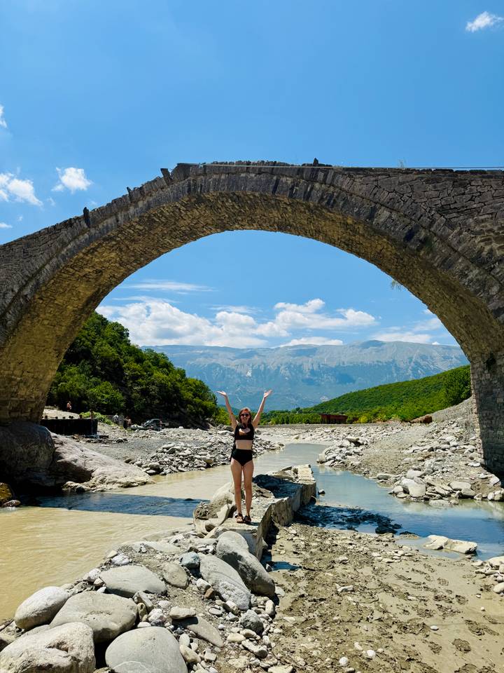 Woman in swimwear celebrating beneath a massive stone arch bridge spanning a rocky riverbed