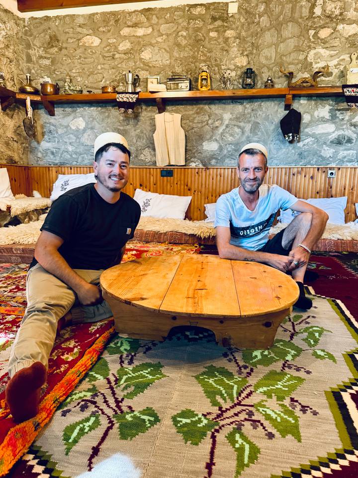 Two men sitting on colorful rugs inside a rustic wood-paneled guesthouse around a low round table