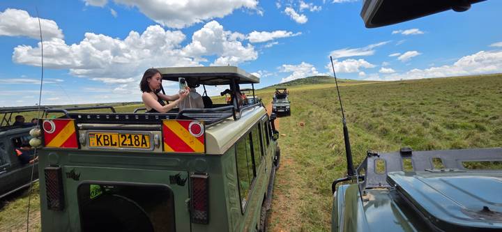 Tourists stand in open-top safari jeeps photographing wildlife on vast grass plains.