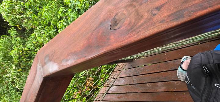 Wooden railing of a treetop walkway with a backpack and part of a shoe in view against green foliage.