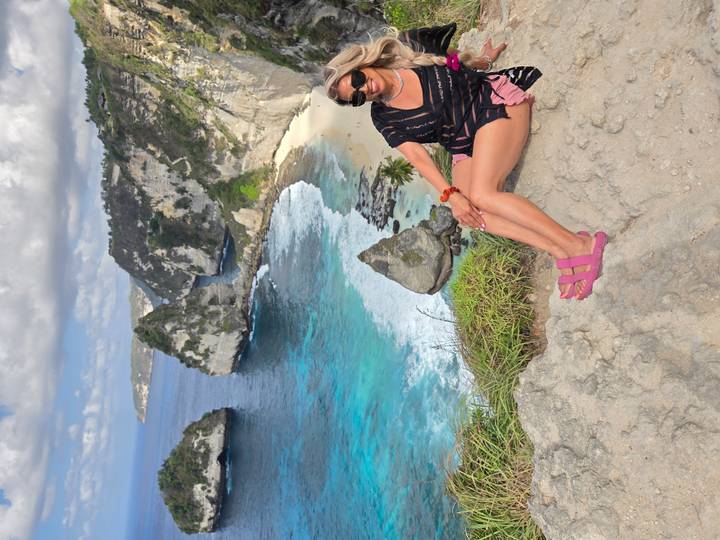 Woman seated on a cliff edge smiles with dramatic coastal limestone cliffs and turquoise bay behind.