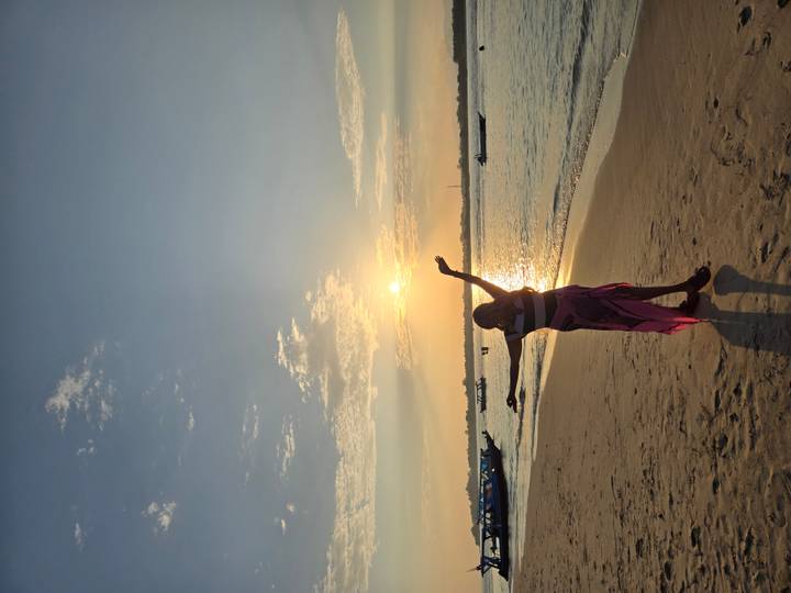 Silhouette of a woman on a sandy beach at sunset with boats on the water.