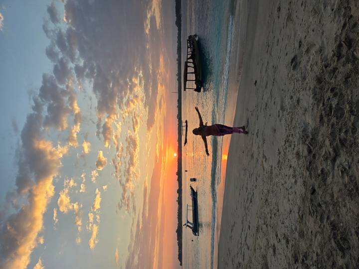A woman with outstretched arms stands on the shore as the sun rises behind distant boats.