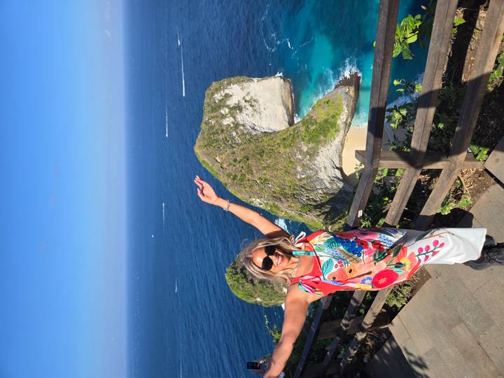 Woman raises arms at the famous Kelingking viewpoint overlooking dramatic cliffs and blue sea.