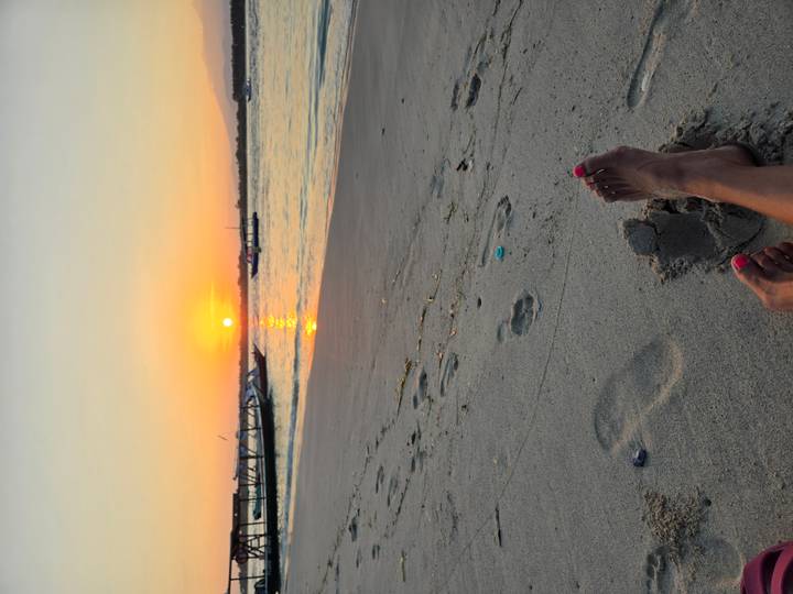 View of sandy beach and boats at sunset, with person's feet and pink toenails in foreground.