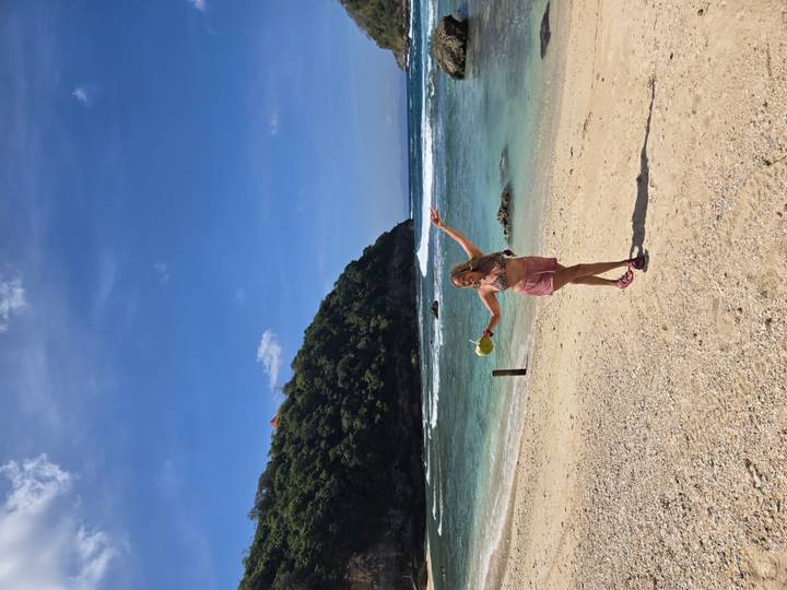 A young woman holding a fresh coconut poses joyfully on a quiet tropical beach with turquoise water and a lush cliff in the background.