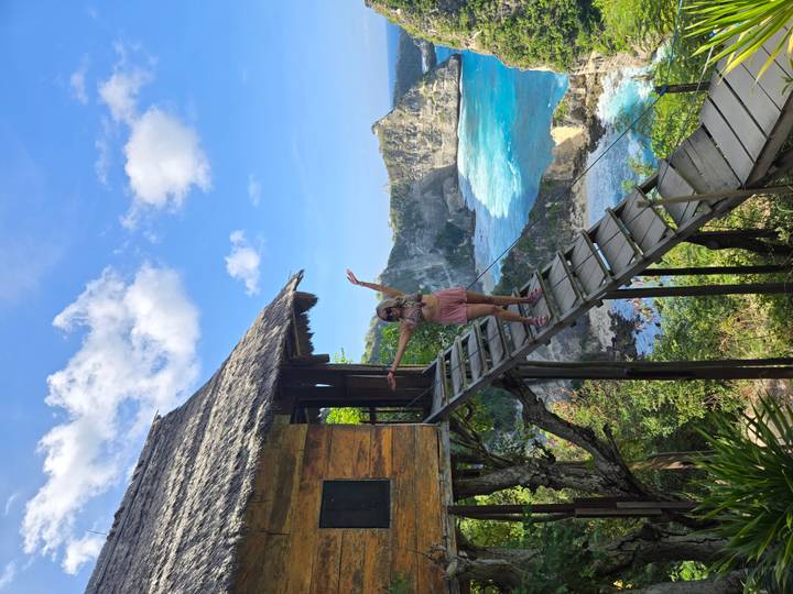 A traveler waves from a wooden treehouse perched high above dramatic cliffs and bright blue sea.