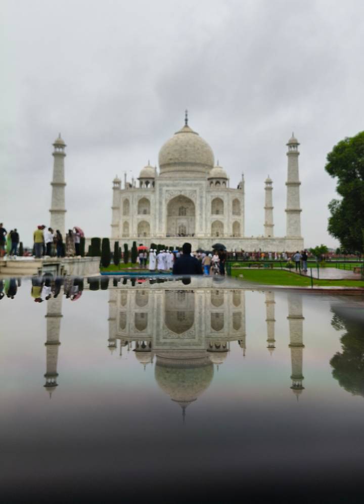 The Taj Mahal reflected in a wet surface on a cloudy day with visitors walking in front.