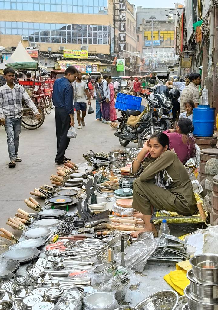A young street vendor sits among stacks of kitchenware on a busy city lane, looking thoughtful.