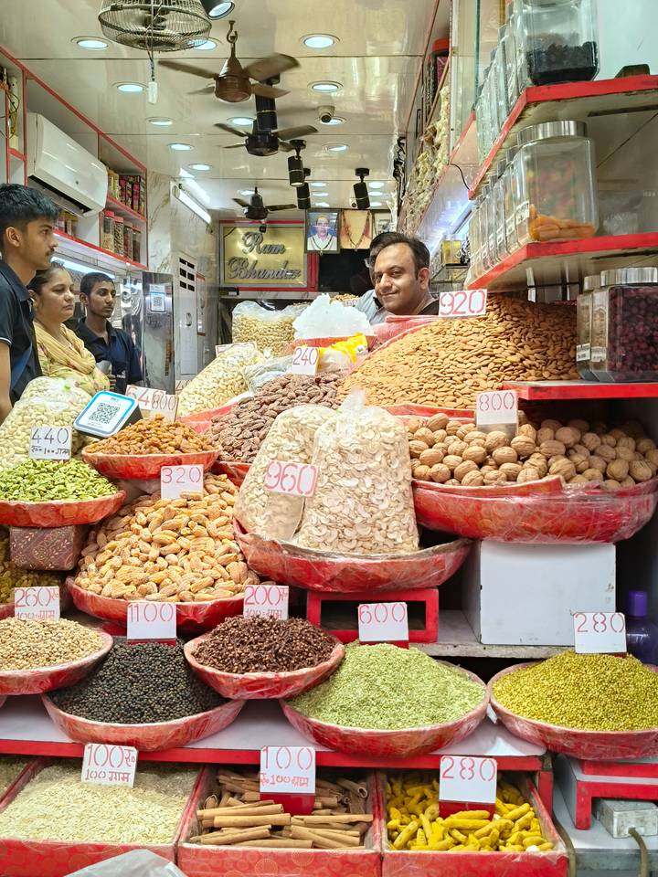 A market stall overflows with nuts and dried fruits labeled with handwritten prices as the vendor smiles.