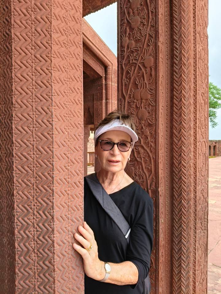 An older woman peeks playfully from behind an ornately carved red sandstone column.