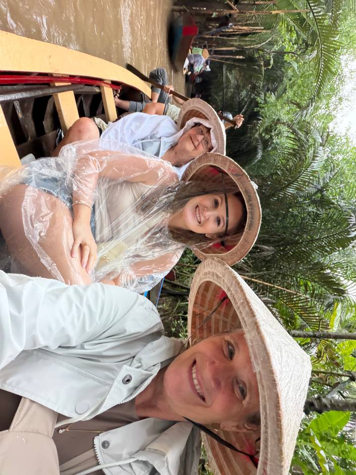 Traveler in poncho and conical hat sits in a wooden sampan amid lush palms.