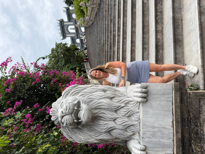 Woman poses beside a stone lion on grand temple steps with bougainvillea overhead.
