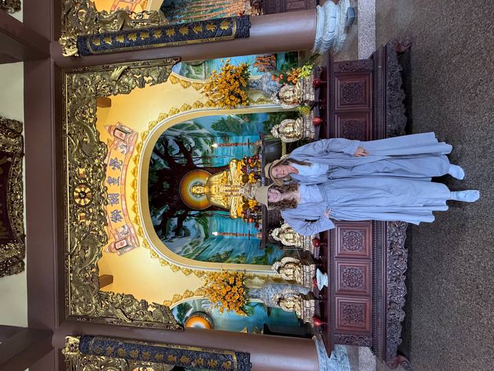 Two women in grey monastic robes stand inside an ornate Buddhist hall.