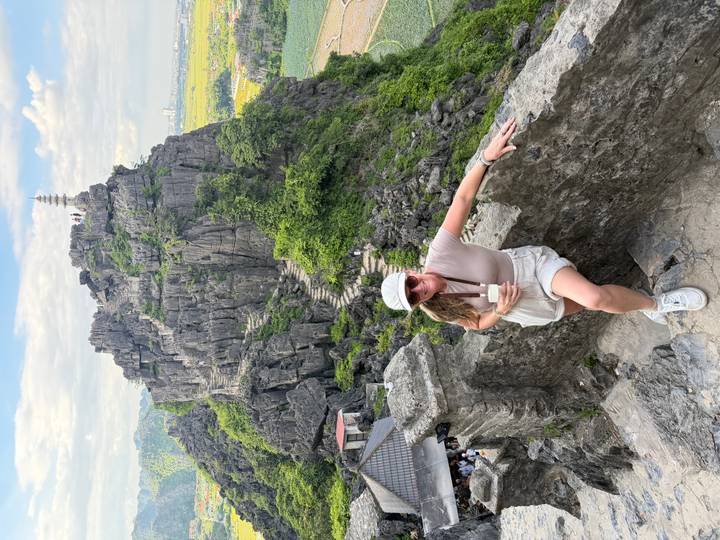 Traveler rests on stone wall with dramatic Mua Cave viewpoint rising behind her.